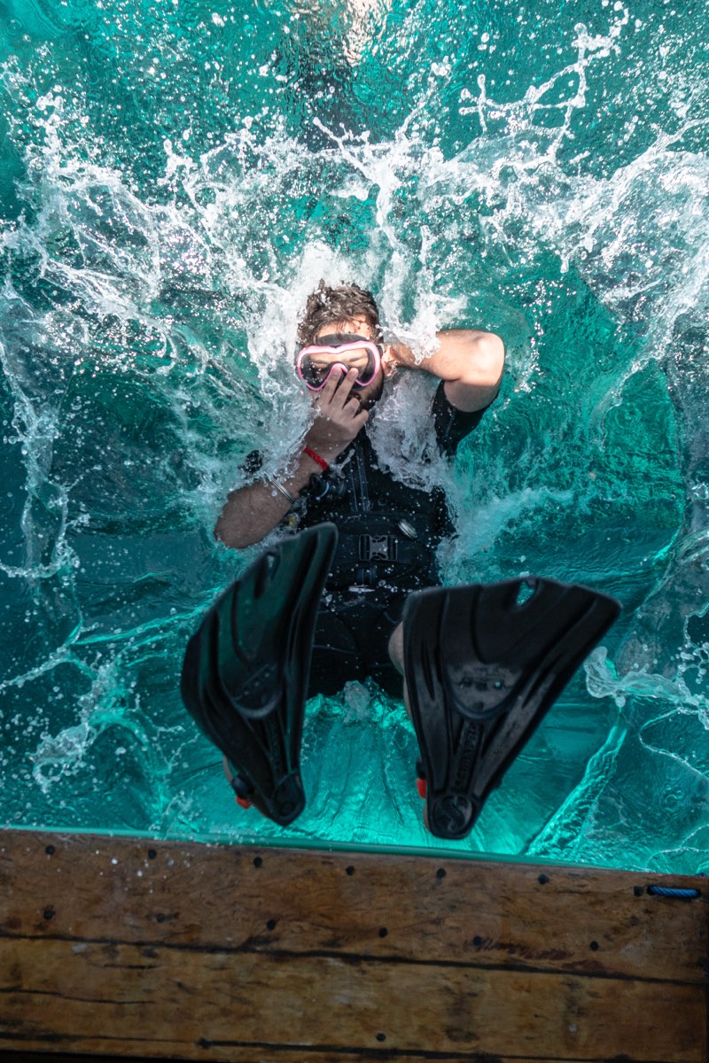 Backroll entry off a dive boat, mid-splash in clear turquoise water, seen from the deck above.